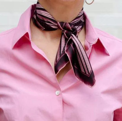 Houston Ruby Silk Bandana from Laslett England with a graphic radial stripe in ruby and rose pink pictured tied at the neck on a female model wearing a pink shirt.