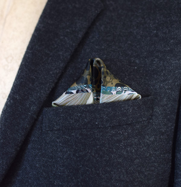 This Marble Camo pocket square applies the flowing, layered quality of traditional hand-marbling to a palette of olive green, teal, black and ivory. Photographed folded into a triangle in a dark suit jacket.
