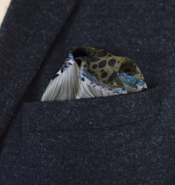 This Marble Camo pocket square applies the flowing, layered quality of traditional hand-marbling to a palette of olive green, teal, black and ivory. Photographed in a dark suit pocket.