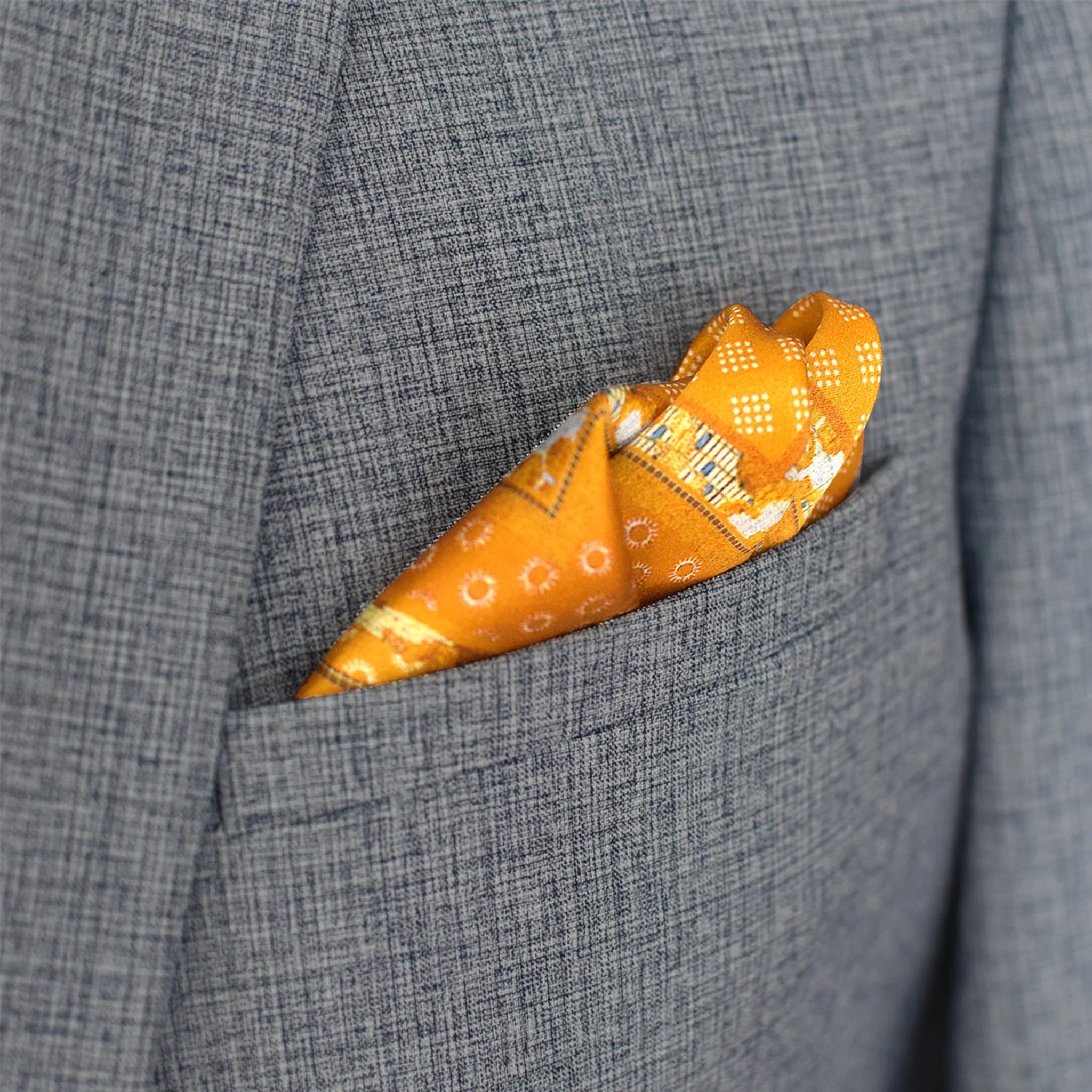 Batik Patchwork Gold pocket square in gold with an intricate patchwork of geometric patterns. Scarf is shown folded in the breast pocket of a grey suit.