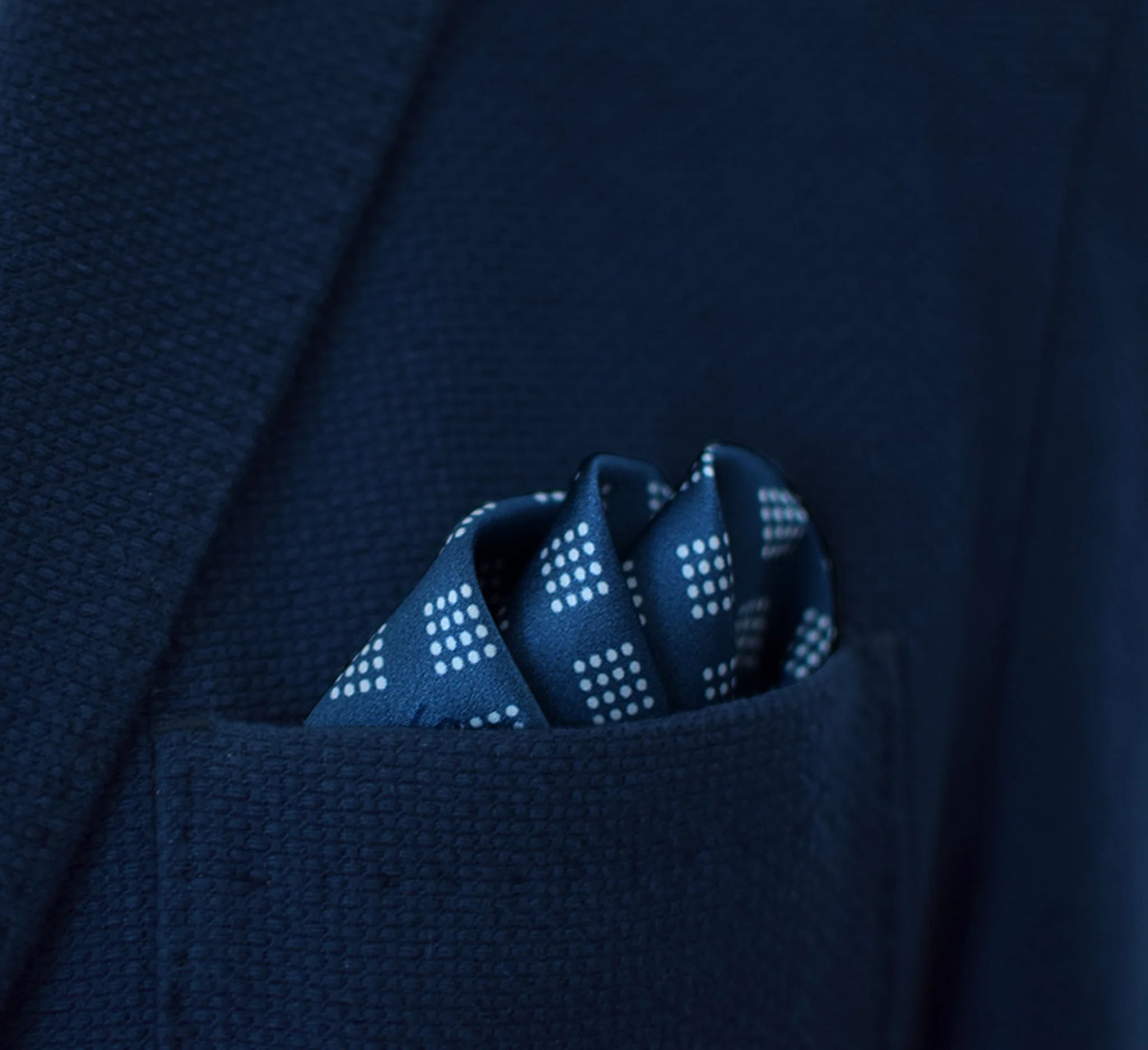Batik Indigo pocket square has a deep ink blue ground with a geometric pattern in the batik tradition, finished with a gingham check border. Square is shown folded into three sections in a blue suit pocket.