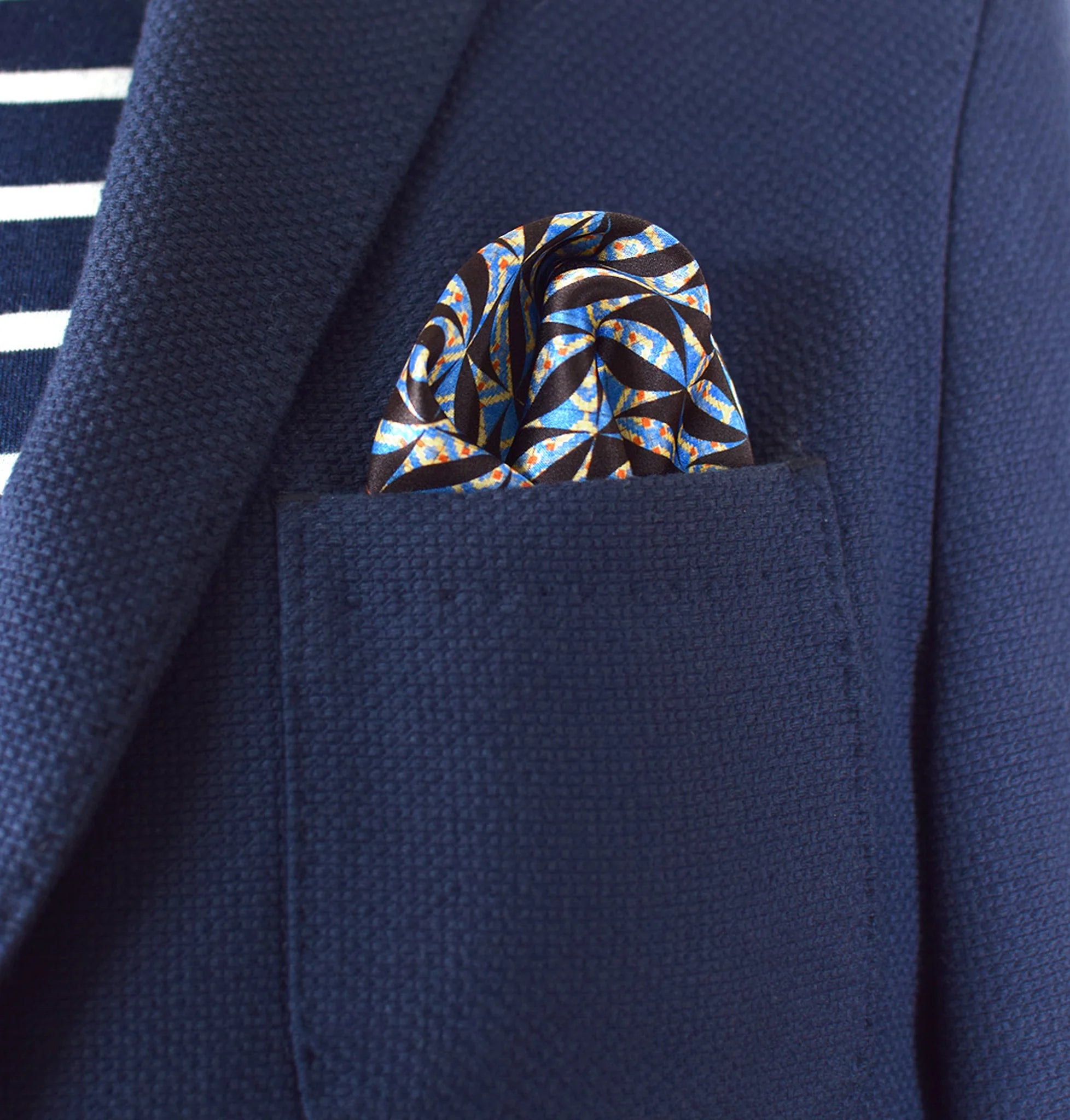 The 100% silk Havana Blue pocket square has a pale blue ground with a diamond repeat carrying small floral motifs in orange, red and cream, finished with a fine geometric border. Square is shown loosely tucked into a blue suit pocket.