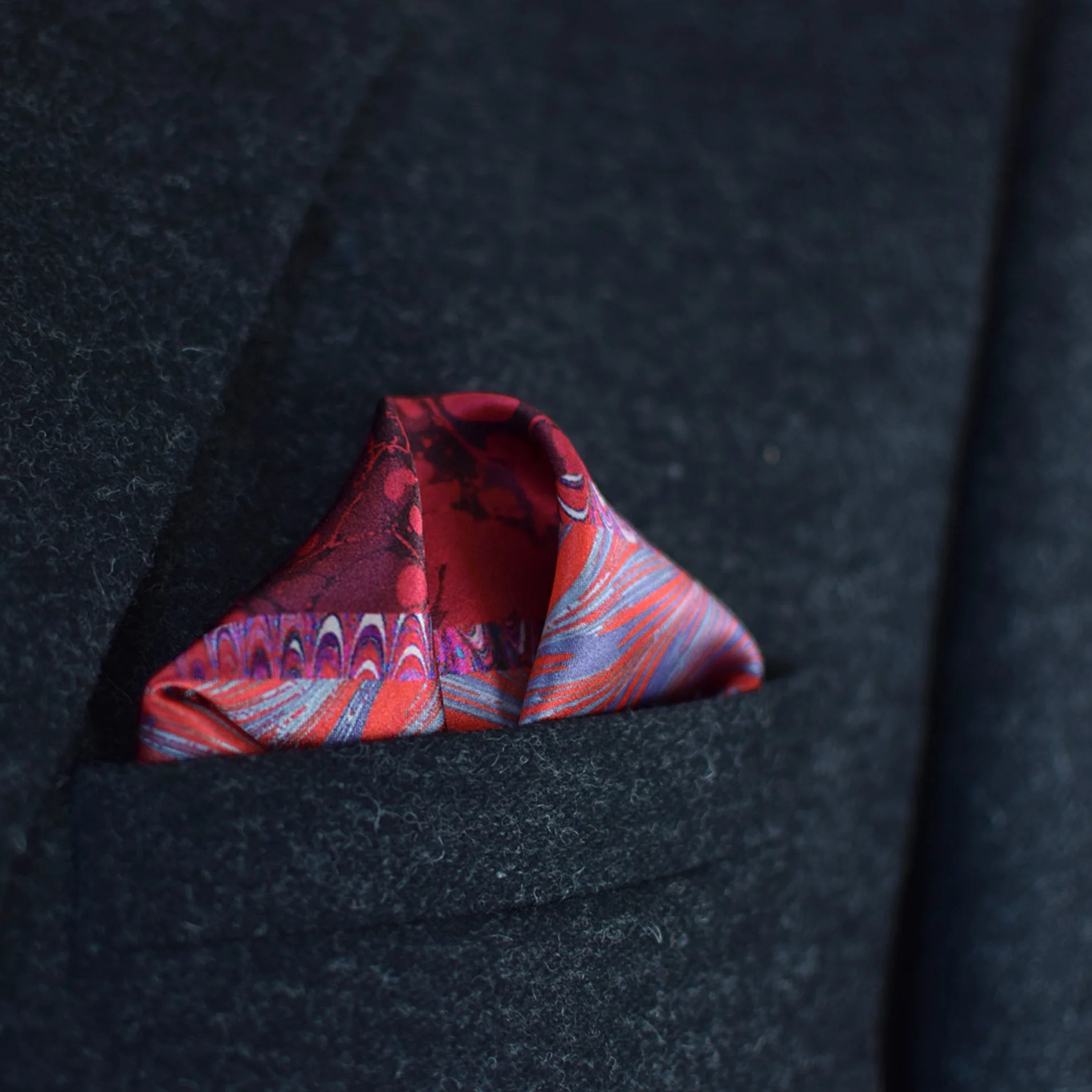 100% silk Marble Red pocket square print carries the flowing, layered quality of traditional hand-marbling in deep red and cream tones with a geometric border. Photographed folded into a triangle in a dark suit pocket.