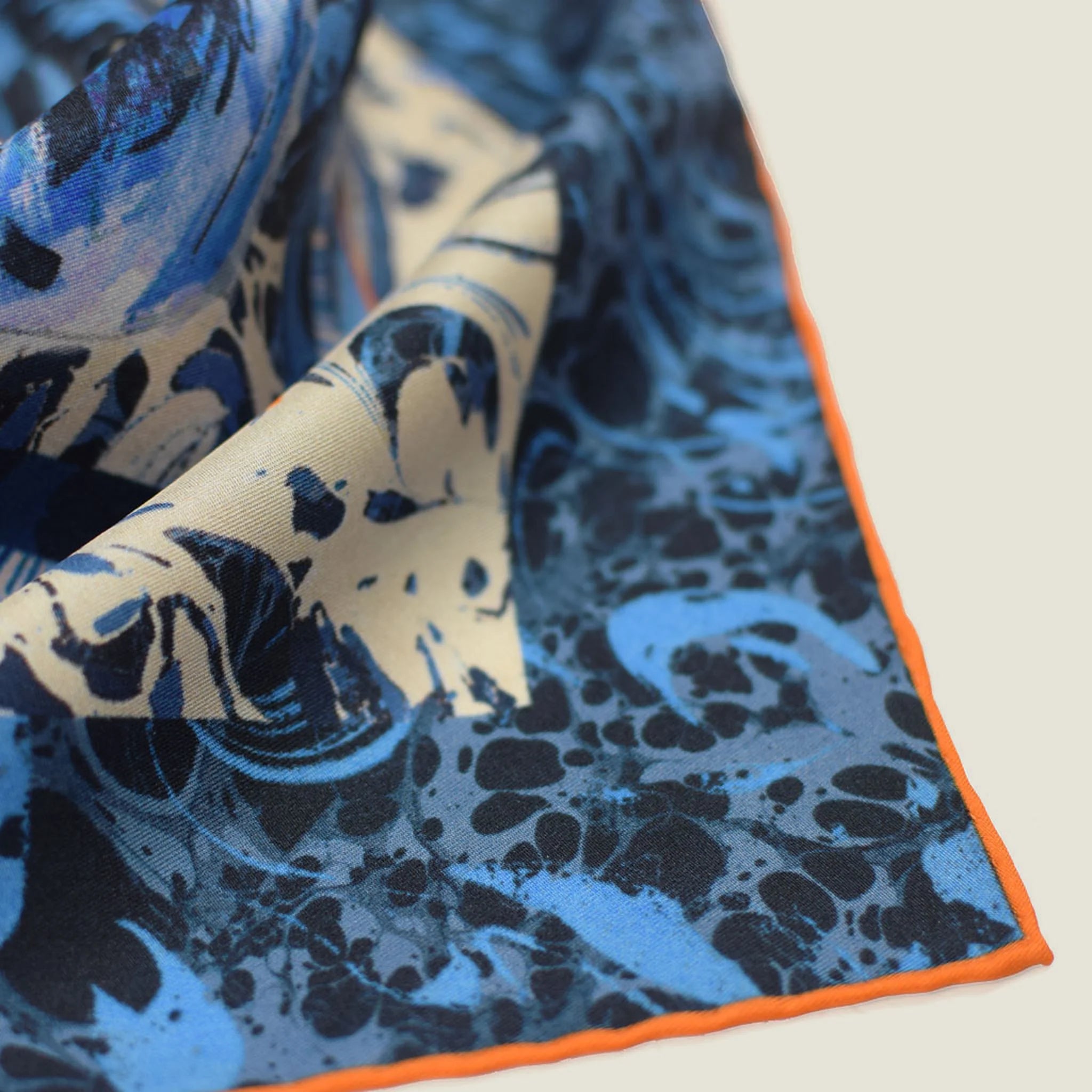 The Laslett England Marble Rose Triangle scarf is shown in a close-up of the triangle edge. Orange piping is shown at the edge and it is laid in slightly rumpled fashion against a neutral background.