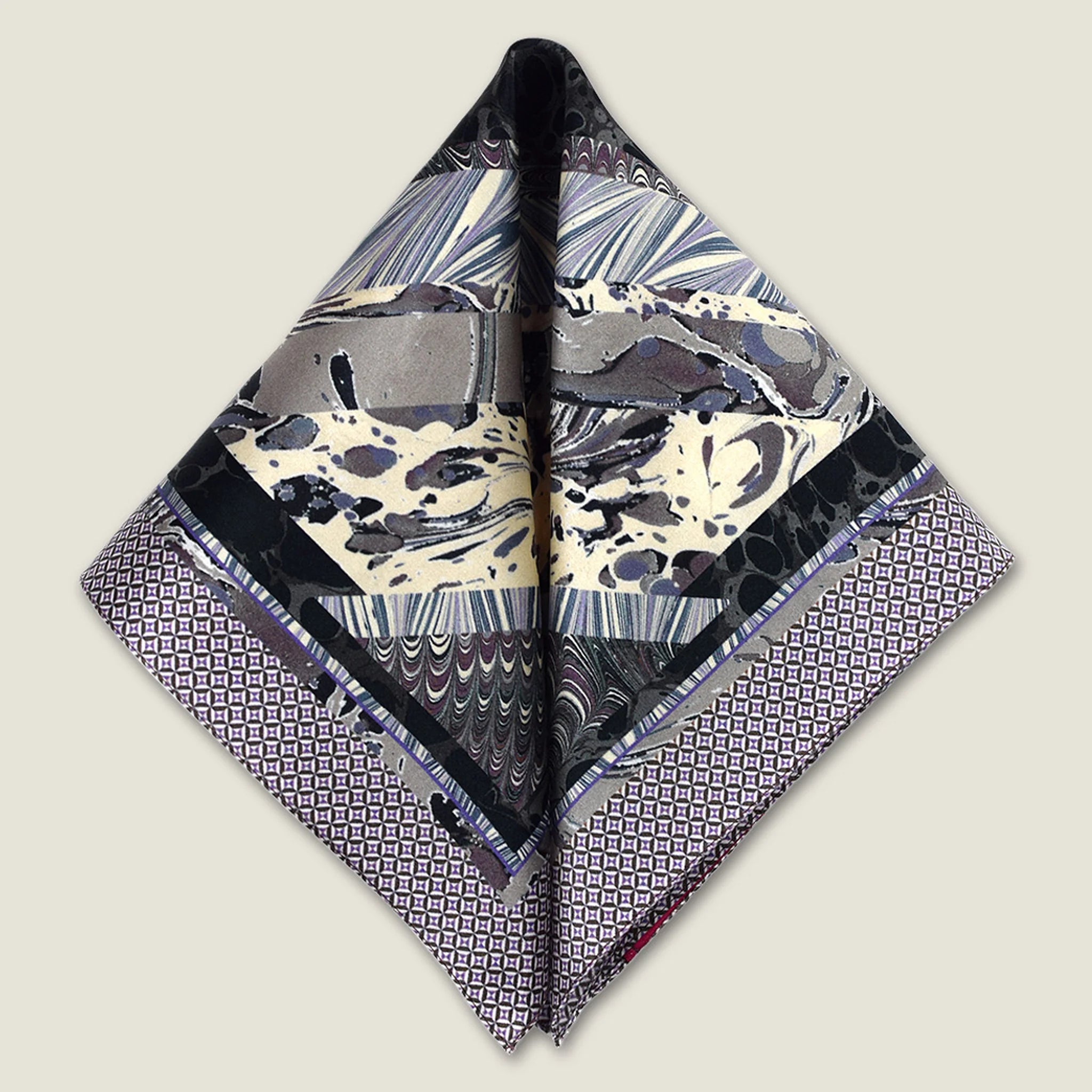 The 100% silk Marble Slate Pocket Square centre field carries multiple marble techniques including feathered combing, stone drop patterns and swirled forms in slate grey, charcoal and dusted lilac, with a geometric tiled border. Photographed folded into two triangles in flat lay.