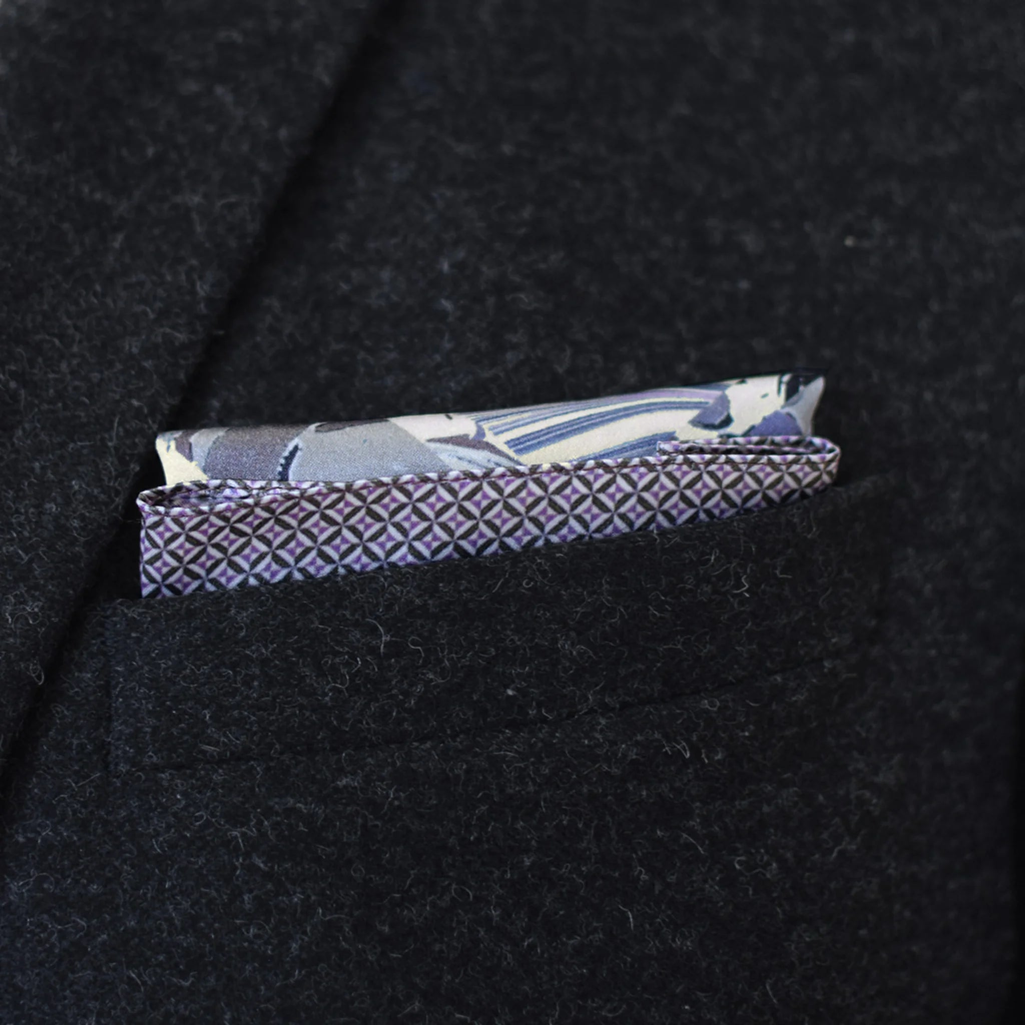 The 100% silk Marble Slate Pocket Square centre field carries multiple marble techniques including feathered combing, stone drop patterns and swirled forms in slate grey, charcoal and dusted lilac, with a geometric tiled border. Photographed folded into a suit jacket in a neat square.
