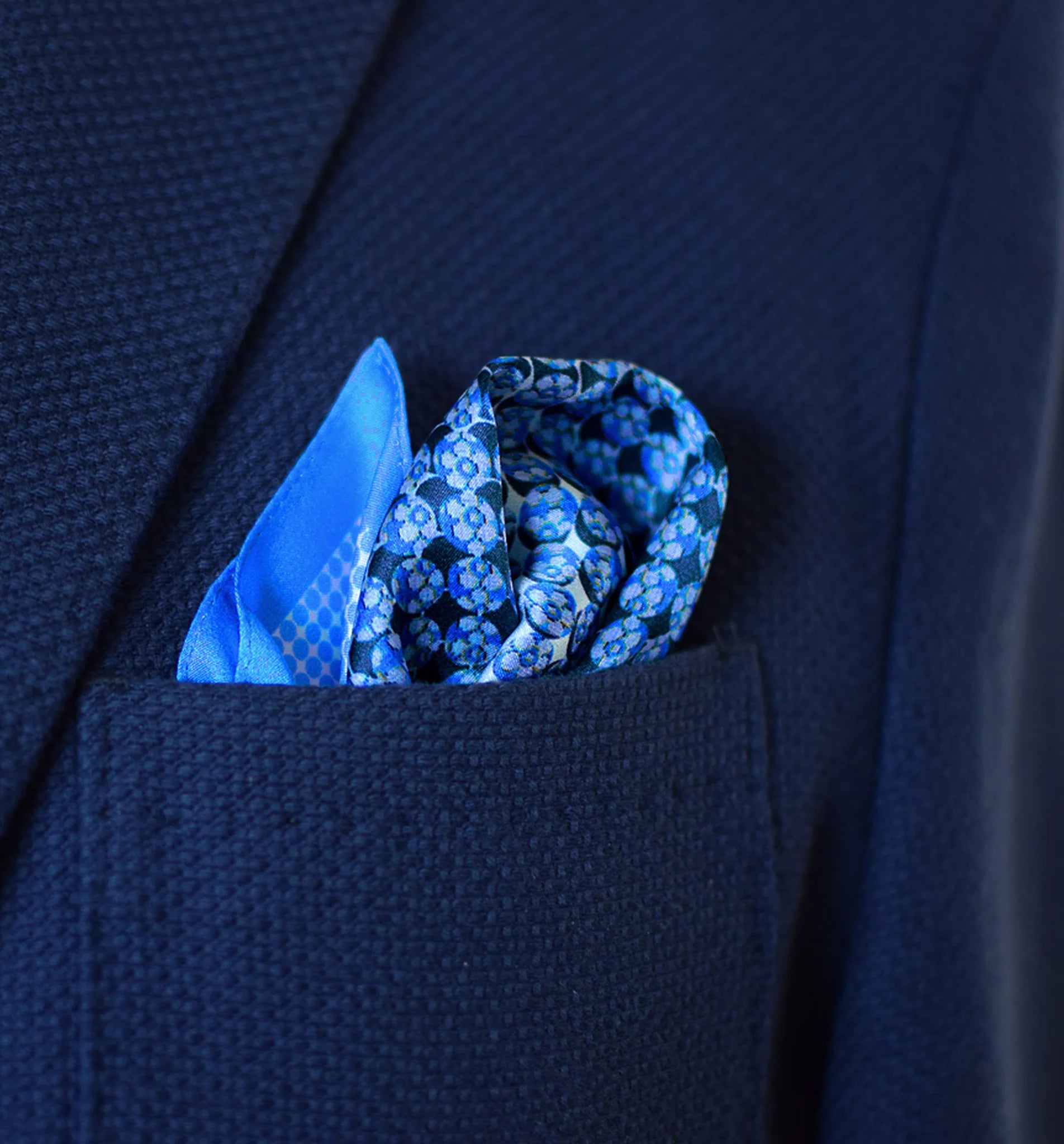 100% silk Matrix Blue pocket square has a distinctive geometric and repeating floral print in blue tones. Shown displayed loosely in a pocket square.