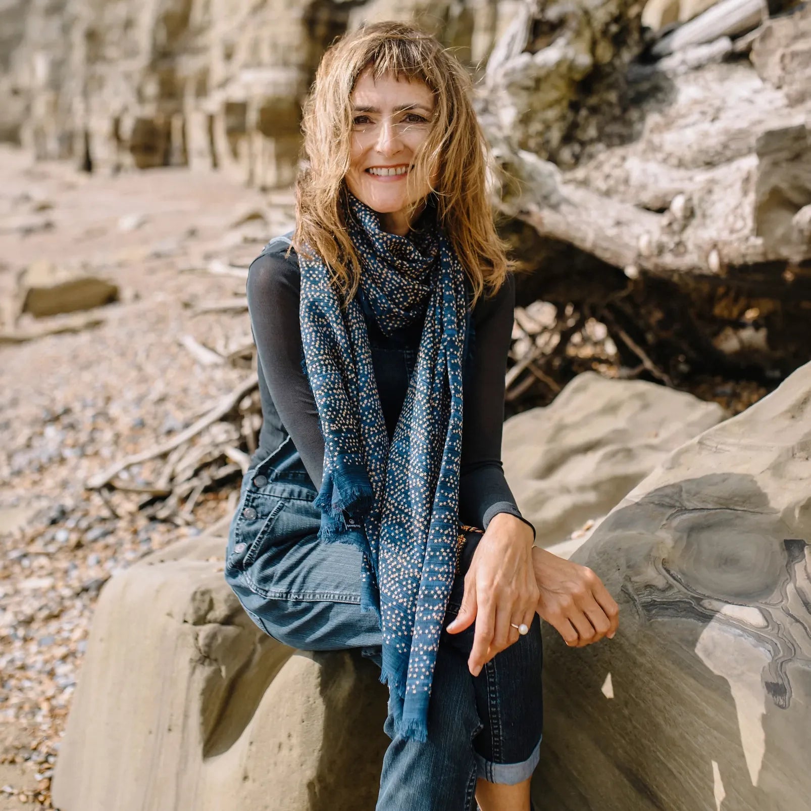Laslett England Starry Indigo Laslett Wrap worn draped by female model seated on rocks on the coast, deep indigo with bronze and gold micro-star print