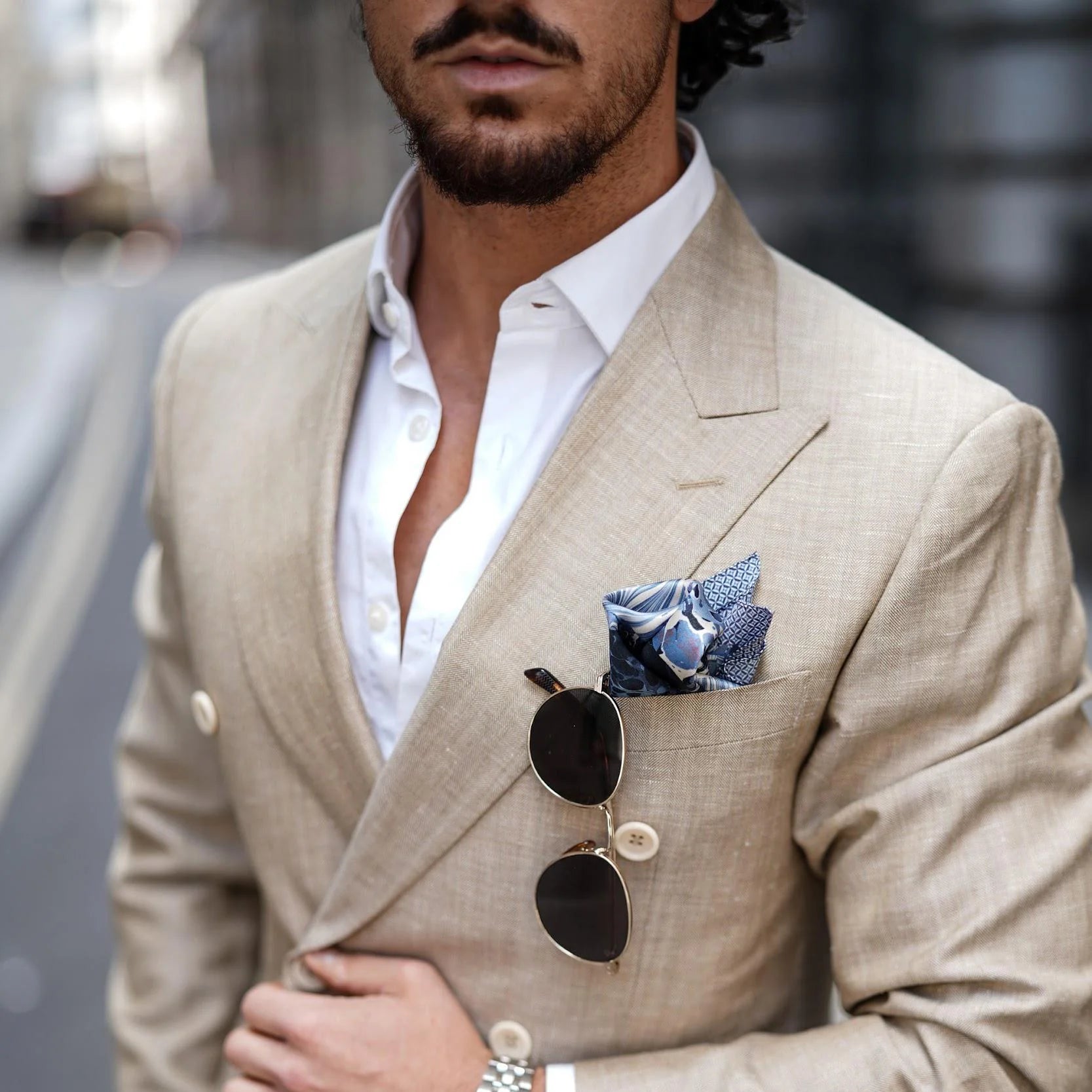 Man wearing a beige suit with sunglasses and a Laslett England Marble Blue pocket square, blurred background