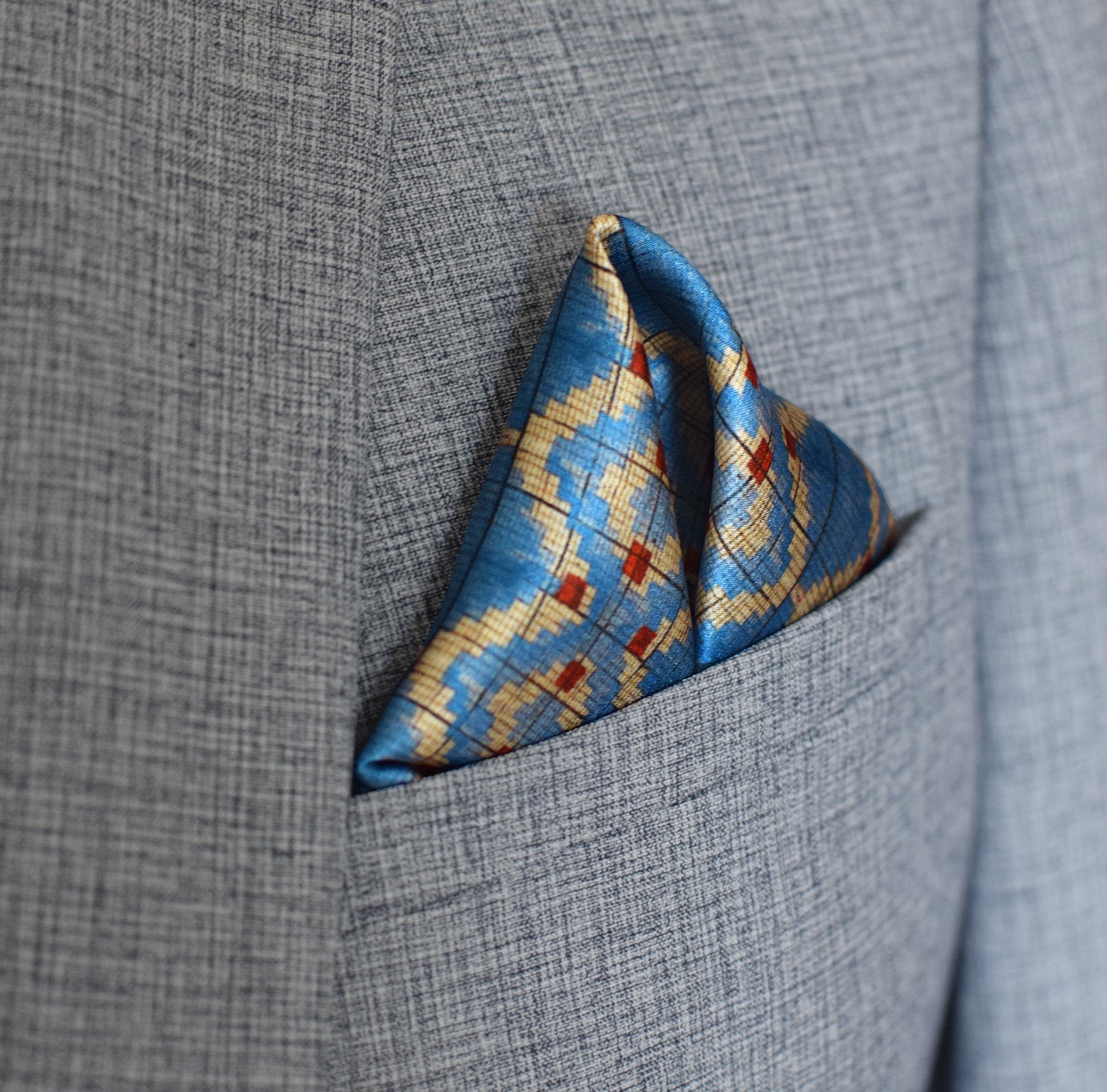 100% silk Aztec Blue Pocket Square in blue by Laslett England. Square is shown folded into a triangle shape in a grey silk suit pocket. 