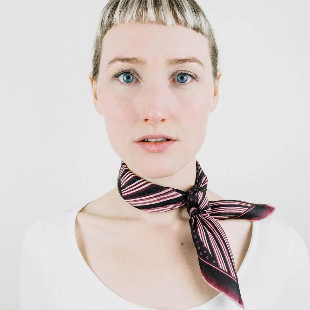 Houston Ruby Silk Bandana from Laslett England with a graphic radial stripe in ruby and rose pink pictured tied at the neck on a female model. 