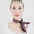 Houston Ruby Silk Bandana from Laslett England with a graphic radial stripe in ruby and rose pink pictured tied at the neck on a female model. 