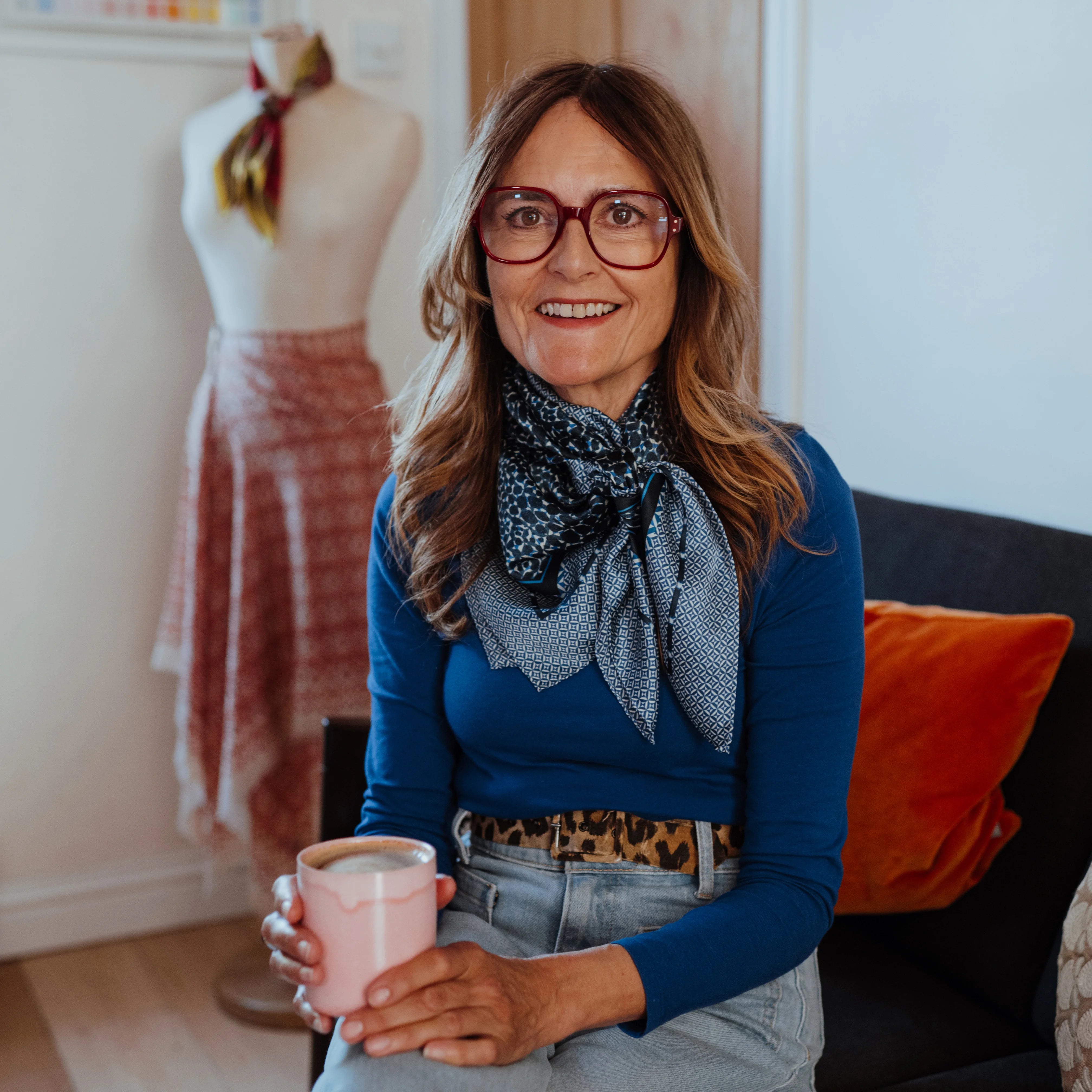 Melanie Laslett wearing the Camo Indigo Classic Square knotted loosely around her neck. She is sitting in her design studio holding a mug of coffee. 