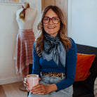 Melanie Laslett wearing the Camo Indigo Classic Square knotted loosely around her neck. She is sitting in her design studio holding a mug of coffee. 