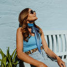 Melanie Laslett is photographed relaxing in sunglasses on a sunny day. She is wearing the Laslett Diamond in Cruise Cobalt wrapped once around the neck adn then tied in a bow. 