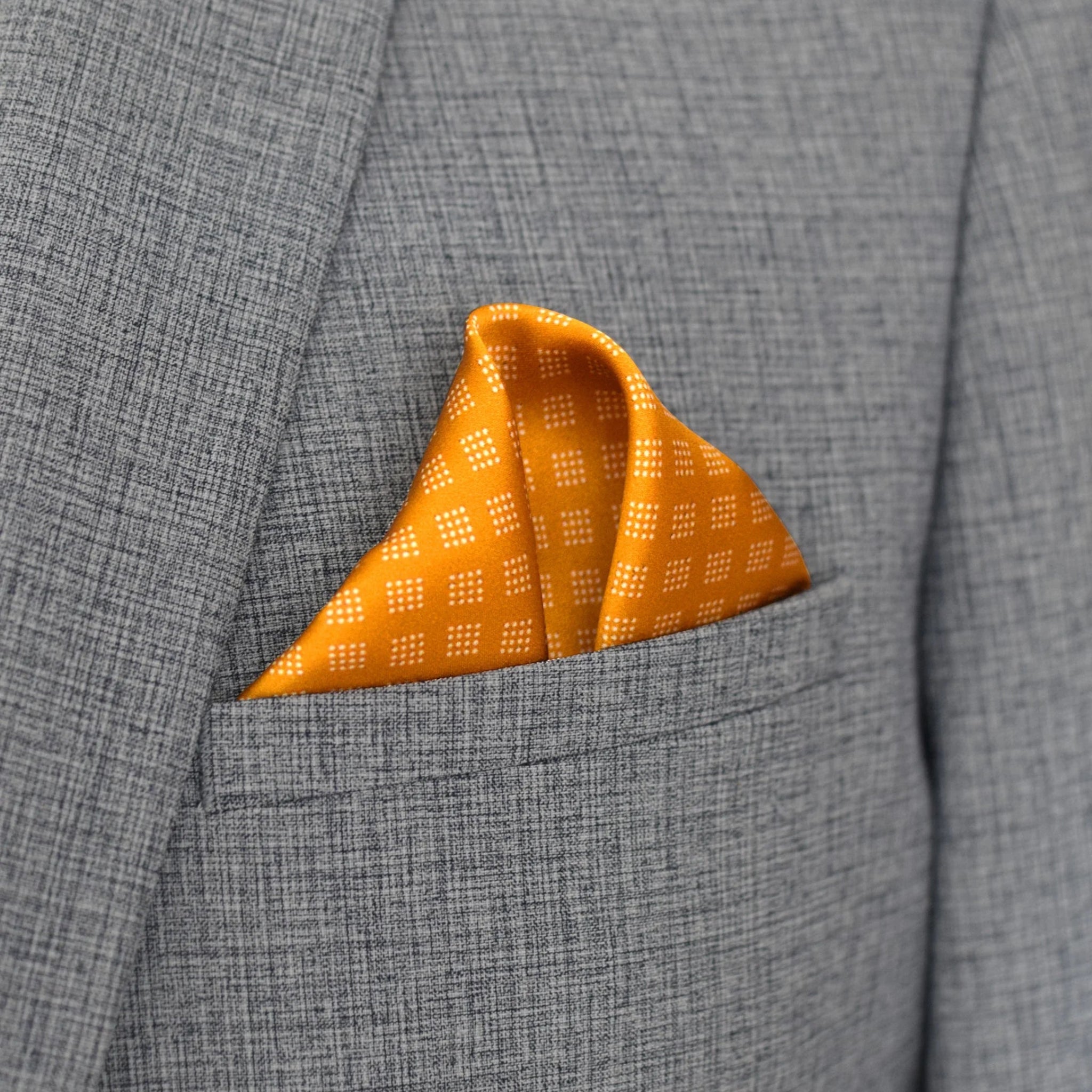 Batik Patchwork Gold pocket square in gold with an intricate patchwork of geometric patterns. Scarf is shown folded in the breast pocket of a grey suit. 