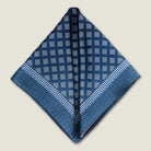 Batik Indigo pocket square has a deep ink blue ground with a geometric pattern in the batik tradition, finished with a gingham check border. Square is shown folded into two triangles against a neutral background. 