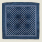 Batik Indigo pocket square has a deep ink blue ground with a geometric pattern in the batik tradition, finished with a gingham check border. Square is shown in flat lay against a neutral background. 