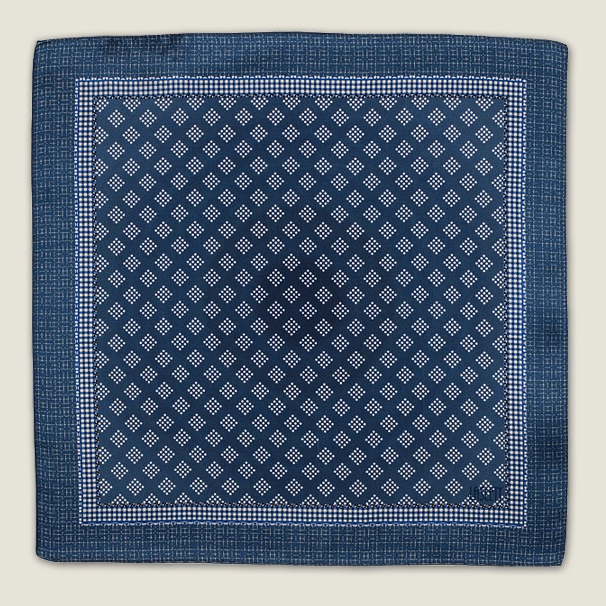 Batik Indigo pocket square has a deep ink blue ground with a geometric pattern in the batik tradition, finished with a gingham check border. Square is shown in flat lay against a neutral background. 