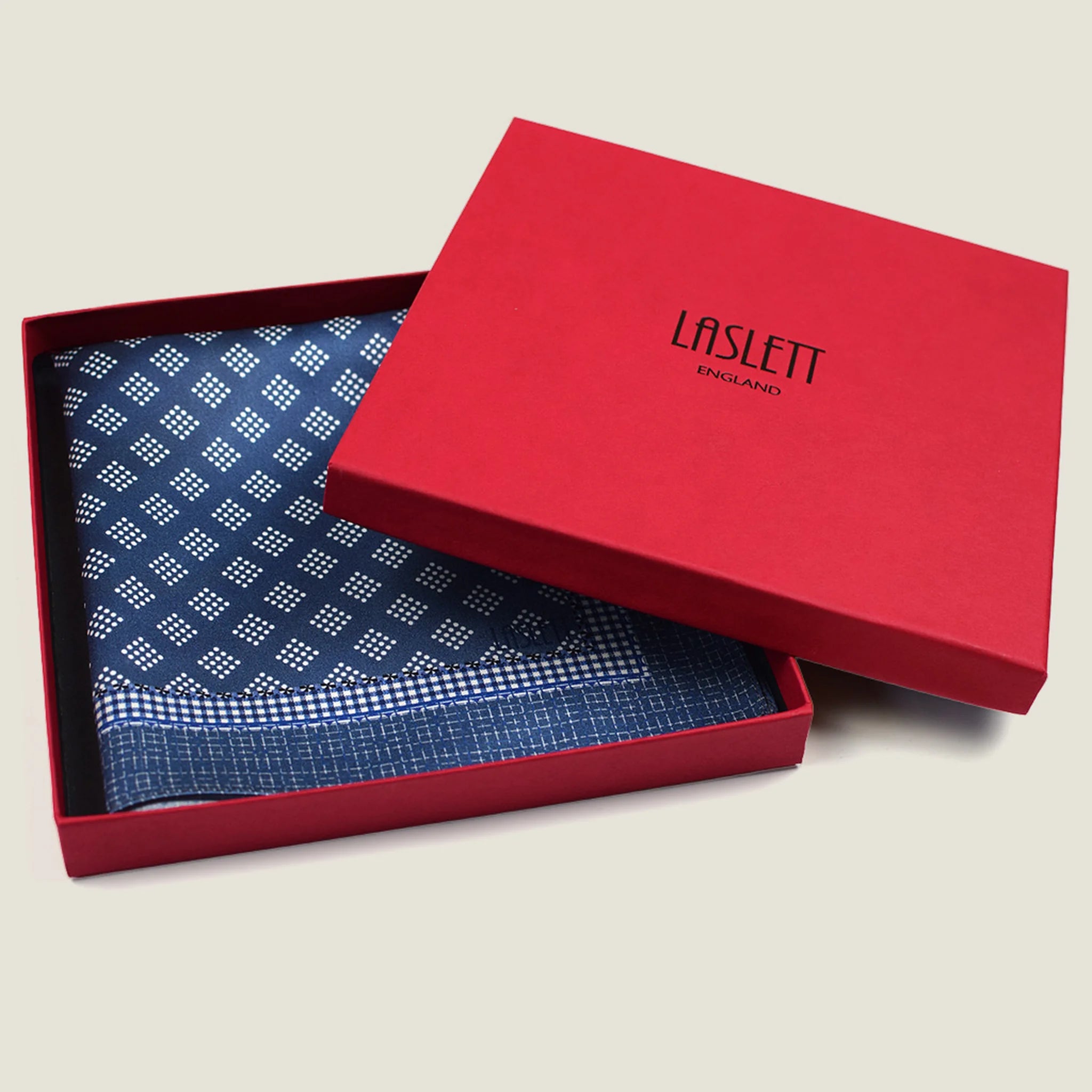 Batik Indigo pocket square has a deep ink blue ground with a geometric pattern in the batik tradition, finished with a gingham check border. Square is shown folded into the red signature Laslett England box. 