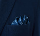 Batik Indigo pocket square has a deep ink blue ground with a geometric pattern in the batik tradition, finished with a gingham check border. Square is shown folded into three sections in a blue suit pocket. 