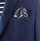 The 100% silk Havana Blue pocket square has a pale blue ground with a diamond repeat carrying small floral motifs in orange, red and cream, finished with a fine geometric border. Square is shown loosely tucked into a blue suit pocket. 