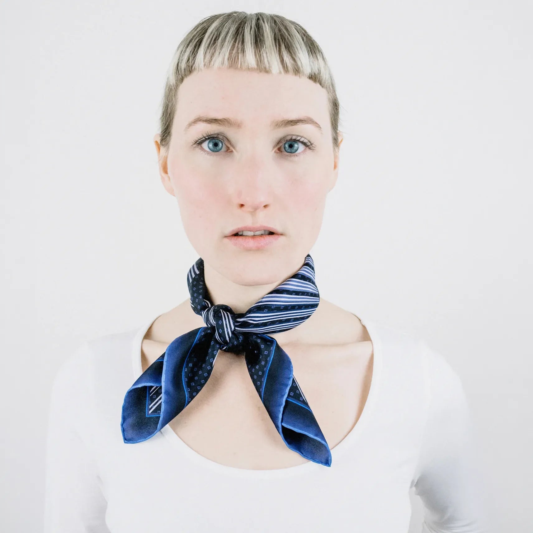 Laslett England Houston Midnight Silk Bandana worn knotted at neck by female model in white top, showing midnight blue graphic radial stripe print on silk twill.