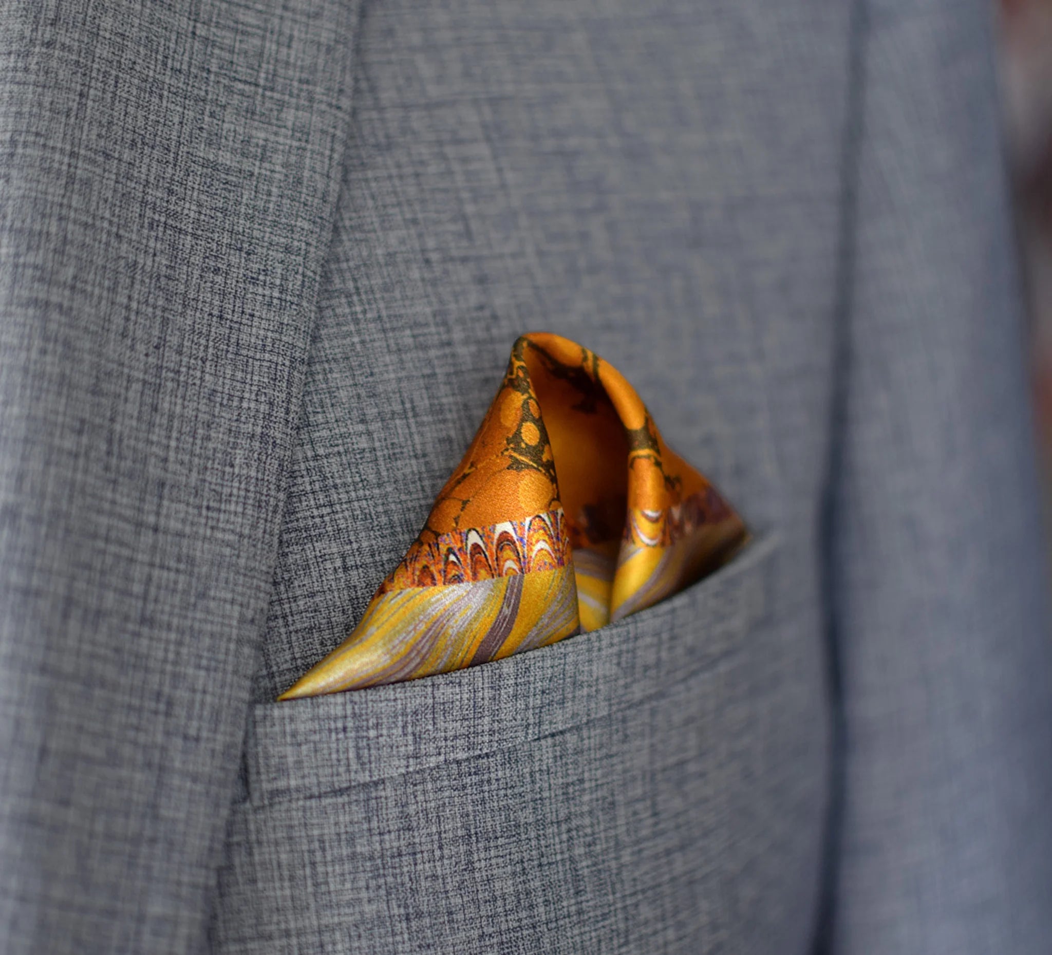 100% silk Marble Amber pocket square is inspired by the decorative marbled papers found inside antique and fine books. Shown tucked neatly in a triangle inside a grey suit jacket. 