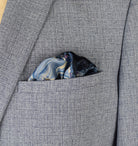 The Marble Blue pocket square print carries the flowing, layered quality of traditional hand-marbling in blue and cream tones with a geometric border. Photographed loosely tucked into a grey suit jacket pocket. 