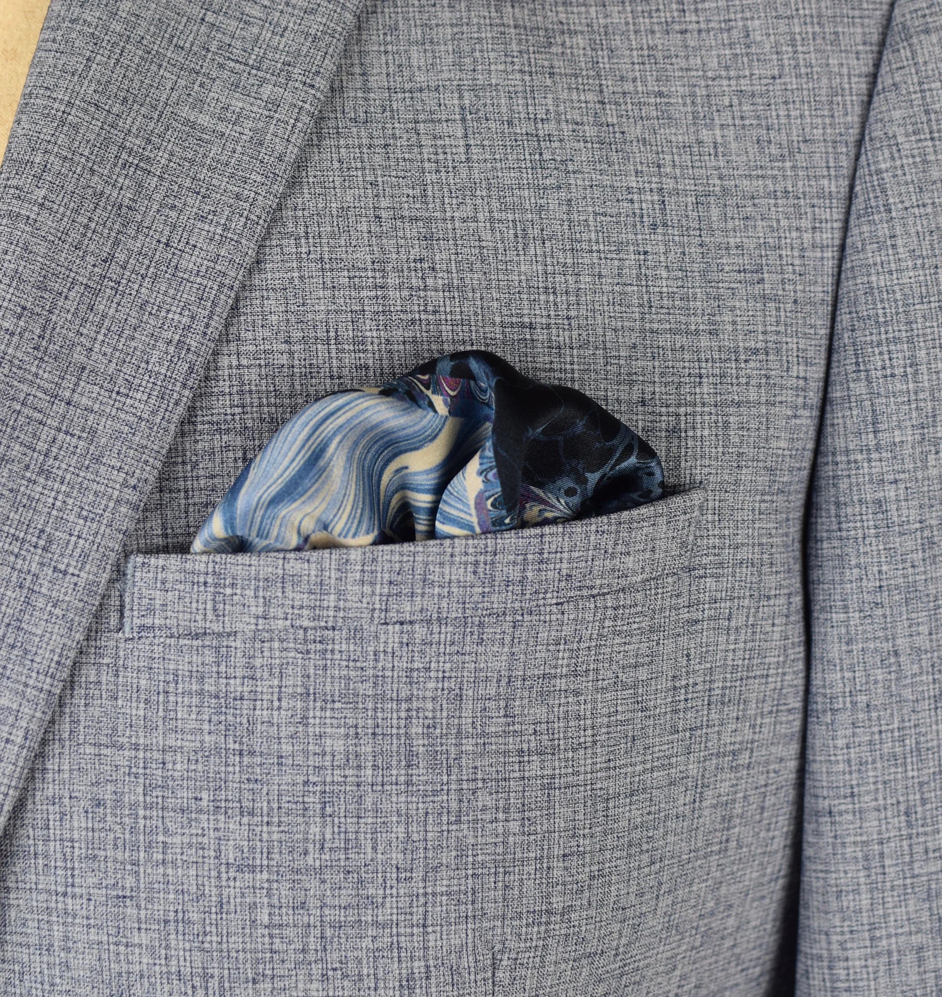 The Marble Blue pocket square print carries the flowing, layered quality of traditional hand-marbling in blue and cream tones with a geometric border. Photographed loosely tucked into a grey suit jacket pocket. 