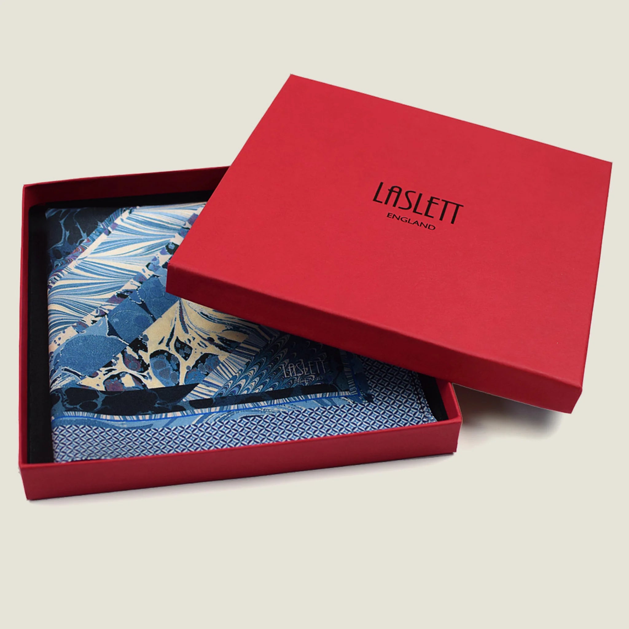 The Marble Blue pocket square print carries the flowing, layered quality of traditional hand-marbling in blue and cream tones with a geometric border. Photographed folded in the red signature box. 
