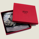 The 100% silk Marble Slate Pocket Square centre field carries multiple marble techniques including feathered combing, stone drop patterns and swirled forms in slate grey, charcoal and dusted lilac, with a geometric tiled border. Photographed in the signature red box. 