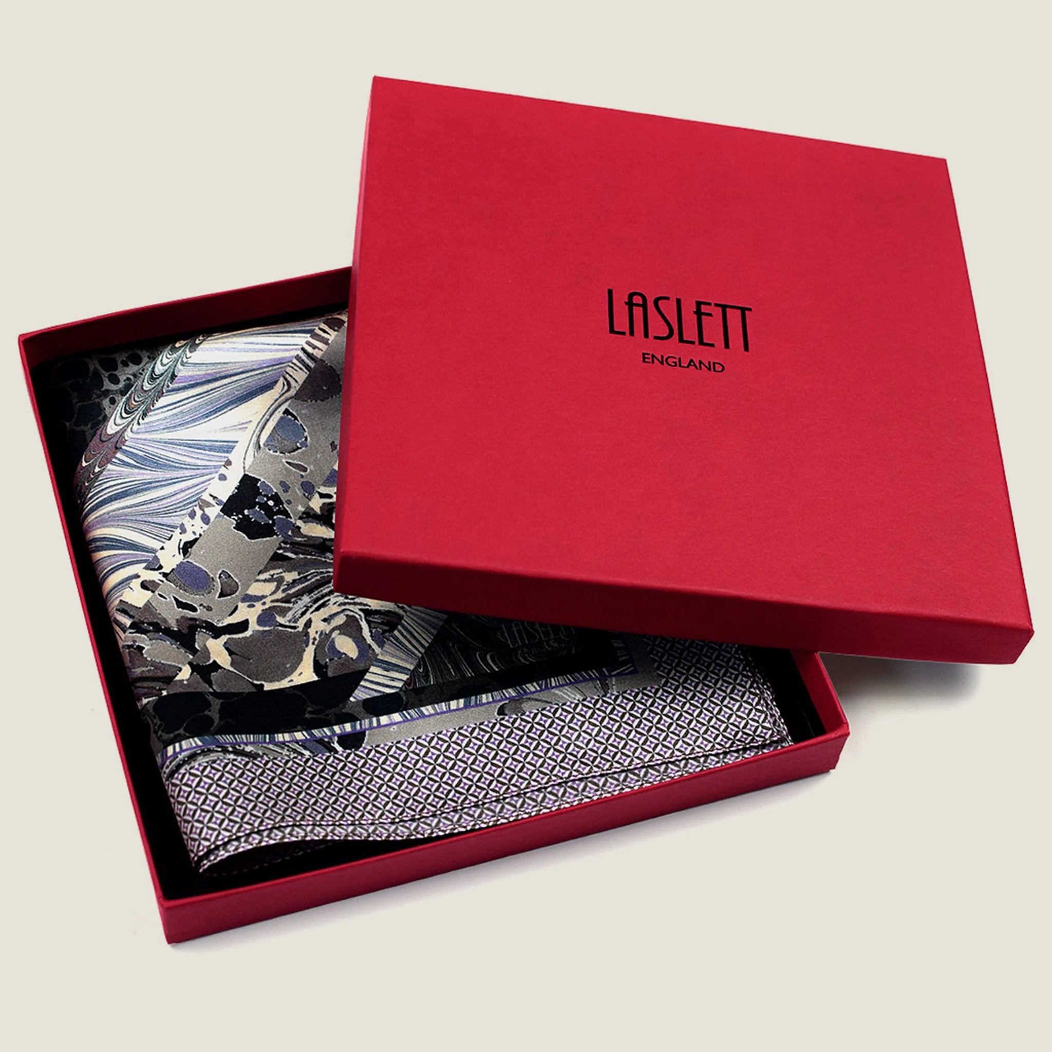 The 100% silk Marble Slate Pocket Square centre field carries multiple marble techniques including feathered combing, stone drop patterns and swirled forms in slate grey, charcoal and dusted lilac, with a geometric tiled border. Photographed in the signature red box. 