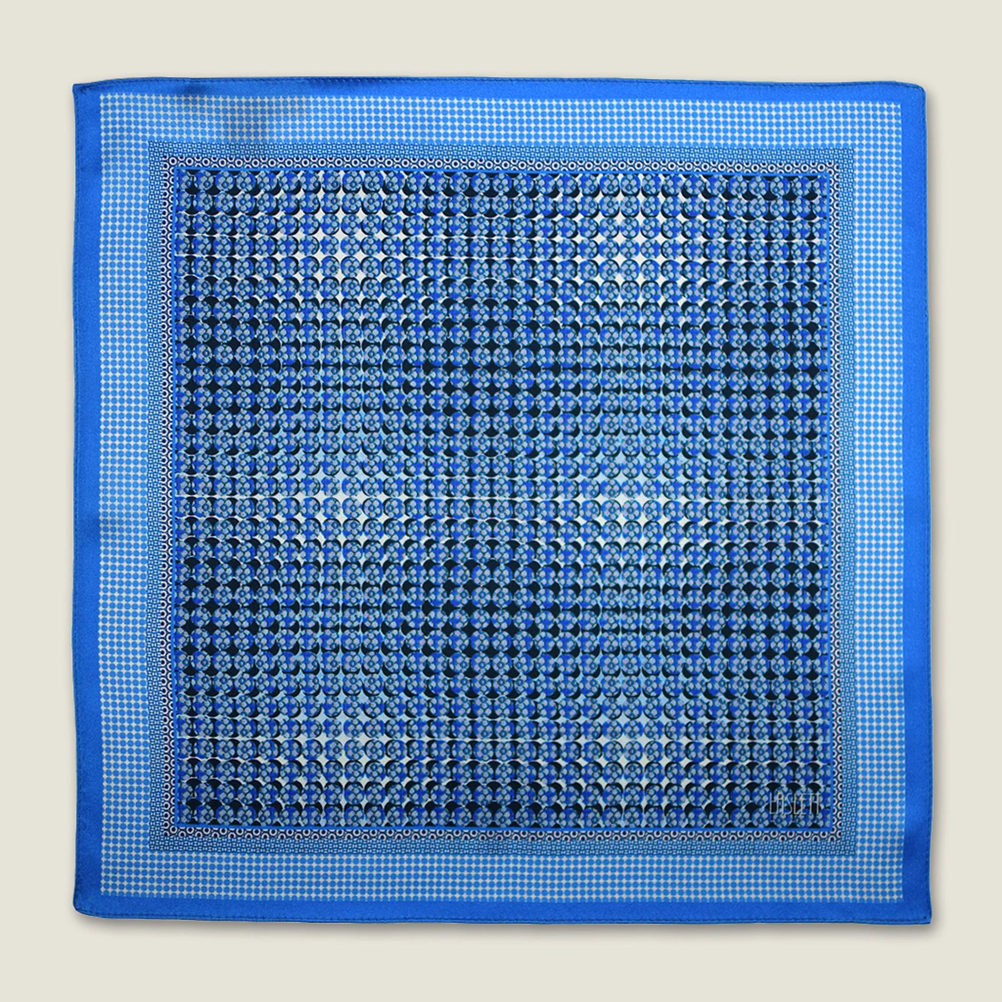 100% silk Matrix Blue pocket square has a distinctive geometric and repeating floral print in blue tones. Shown displayed in flat lay on a neutral background. 