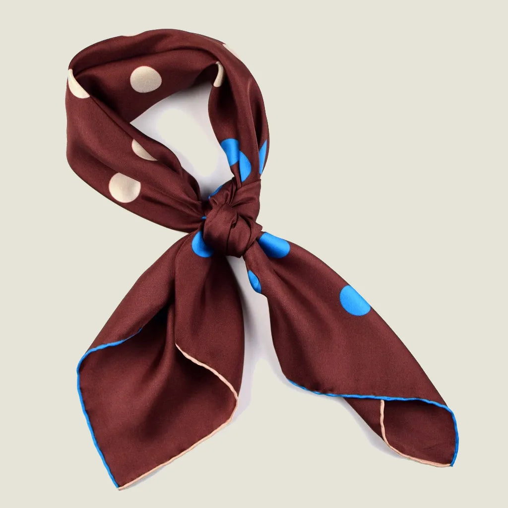 Laslett England Solitaire Chestnut Foulard tied into a knot on a neutral background. Scarf is chestnut silk satin with cornflower blue and cream polka dots.