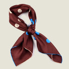 Laslett England Solitaire Chestnut Foulard tied into a knot on a neutral background. Scarf is chestnut silk satin with cornflower blue and cream polka dots.