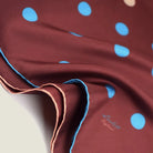 Laslett England Solitaire Chestnut Foulard shown in close up with the signature on display. Scarf is in chestnut silk satin with cornflower blue and cream polka dots.